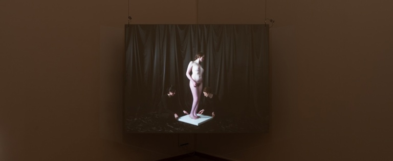 Installationsfoto af Luna Emilie Printz Scales' afgangsværk Eyelids (skærm med nøgen kvinde på plint som drejes af to personer)